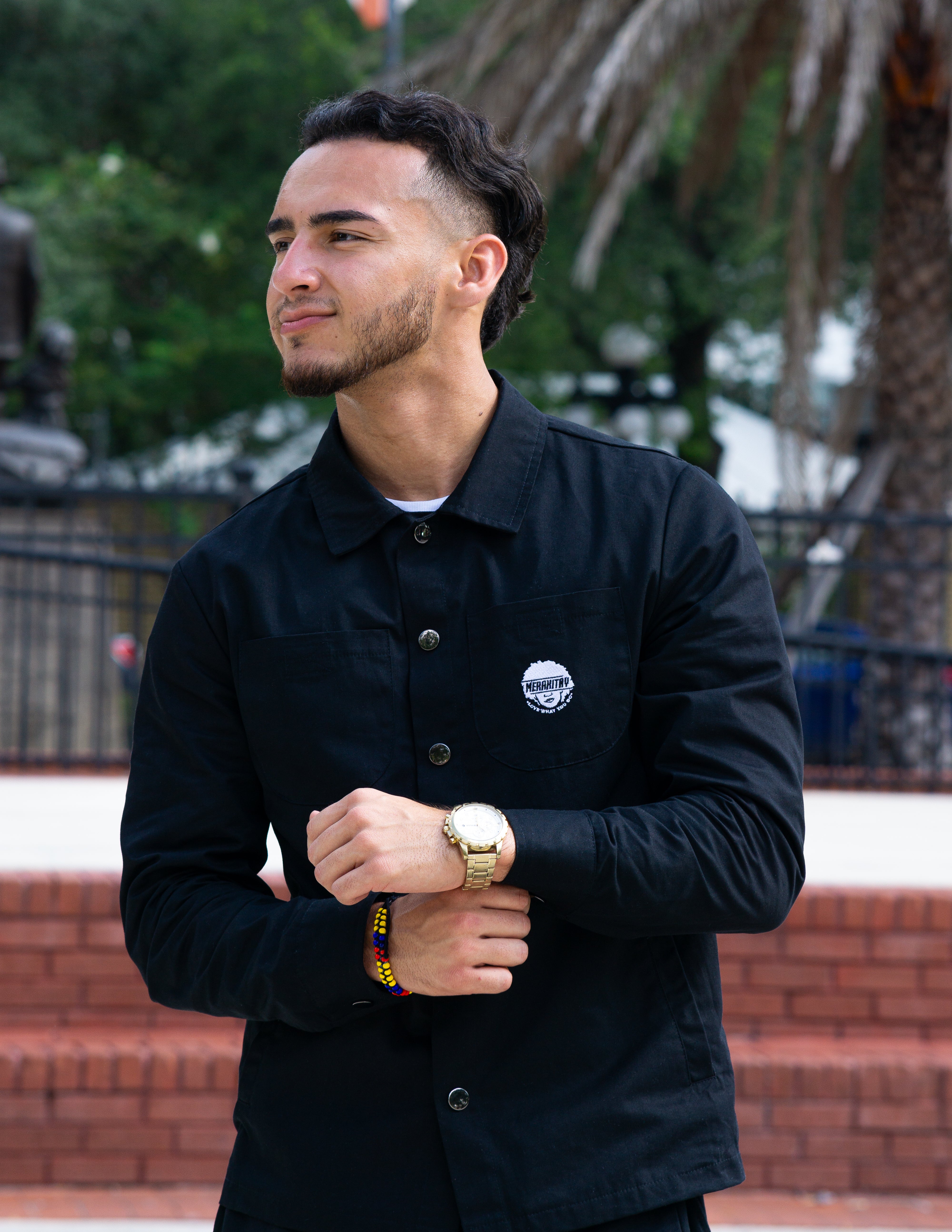 Man wearing the merakitay cargo jacket. Black cargo/ trucker jacket with 4 pockets and buttons for cuffs. Buttons are in the middle of the jacket  to button up if need be. There is a collar as well to be dressed up or dressed down. Embroidered logo. Unisex or gender neutral clothing. 