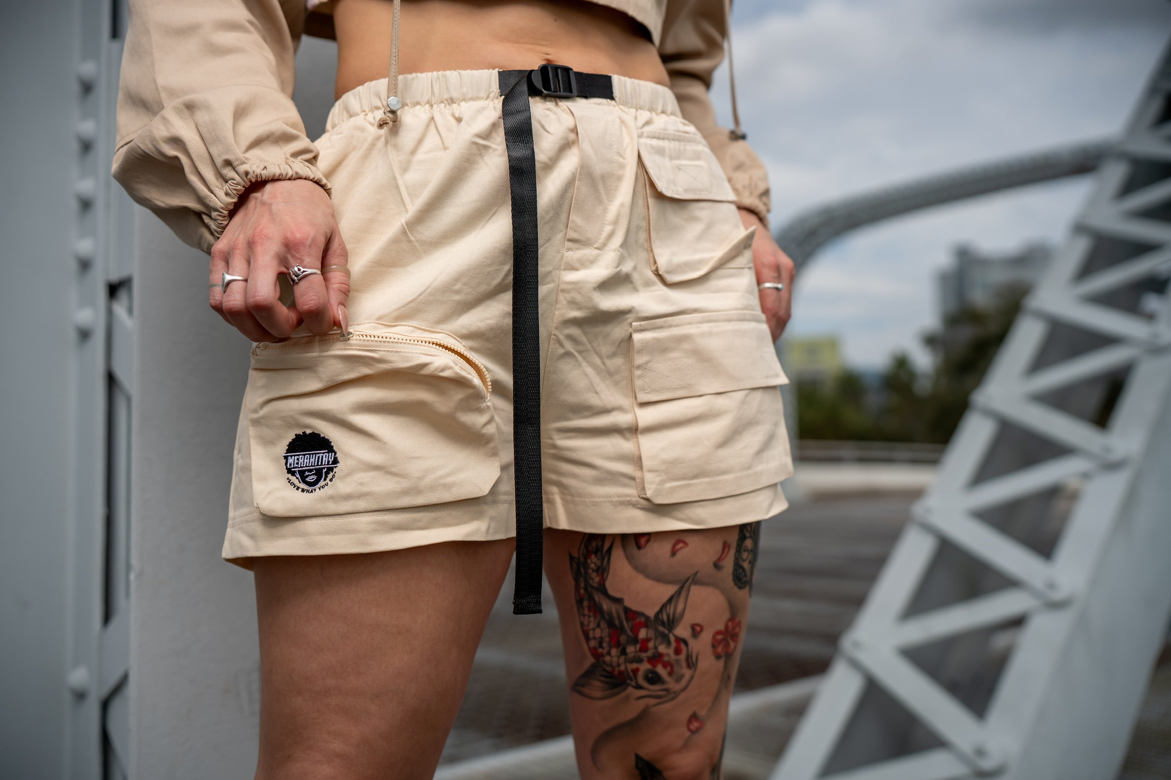 Up close of merakitay cargo shorts. Playing with zipper pocket. Gender neutral cargo shorts. Beige or Cream in color, embroidered logo, adjustable waistband, includes belt, and has six (6) pockets. Mid-thigh length, 3-5 inch inseam.