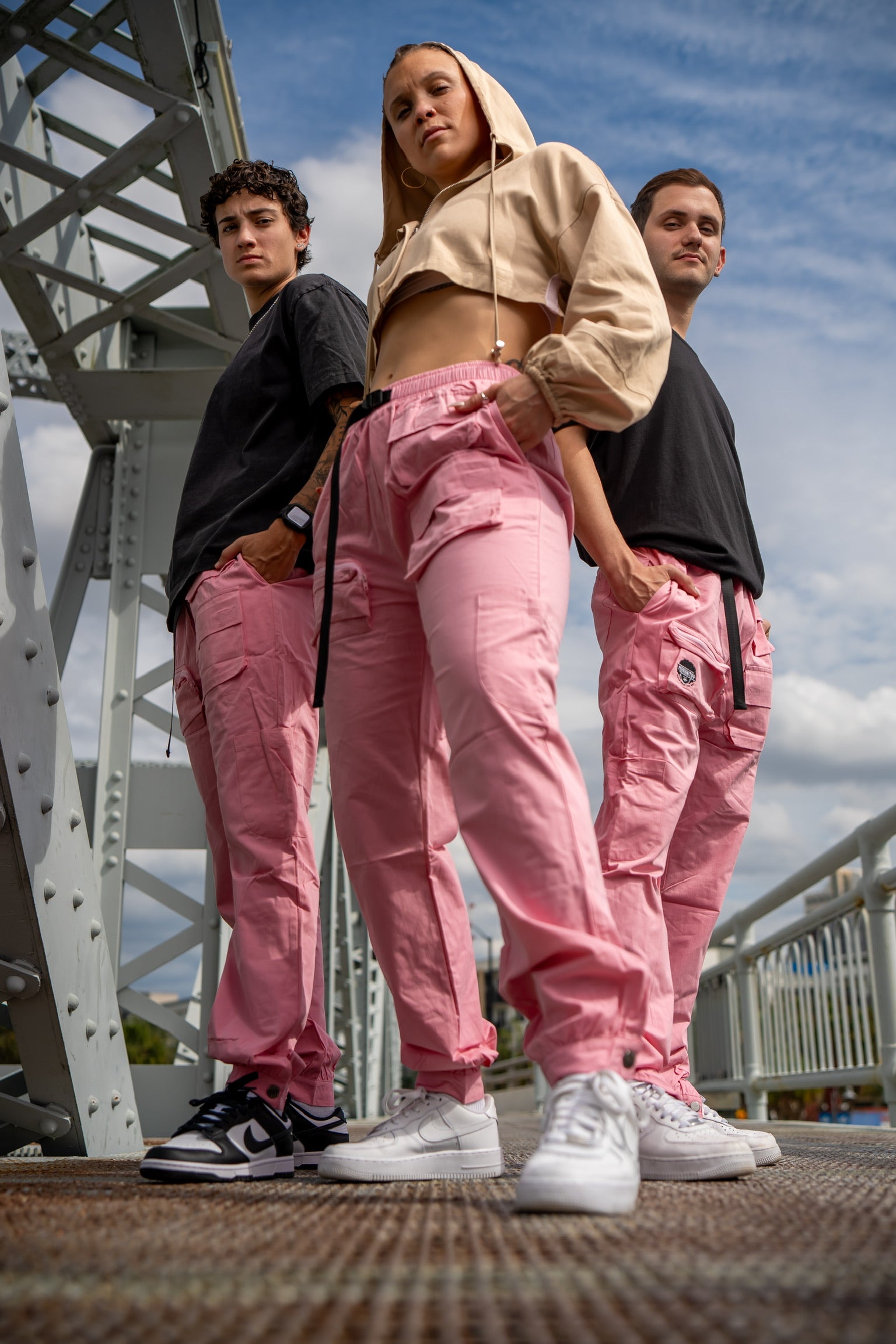 2 women and a man wearing the merakitay pink cargo pants. Pink cargo pants with 9 pockets, stretchy waistband, jogger or straight leg which is adjustable by buttons, embroidered logo, and a belt. Unisex and gender neutral fashion.