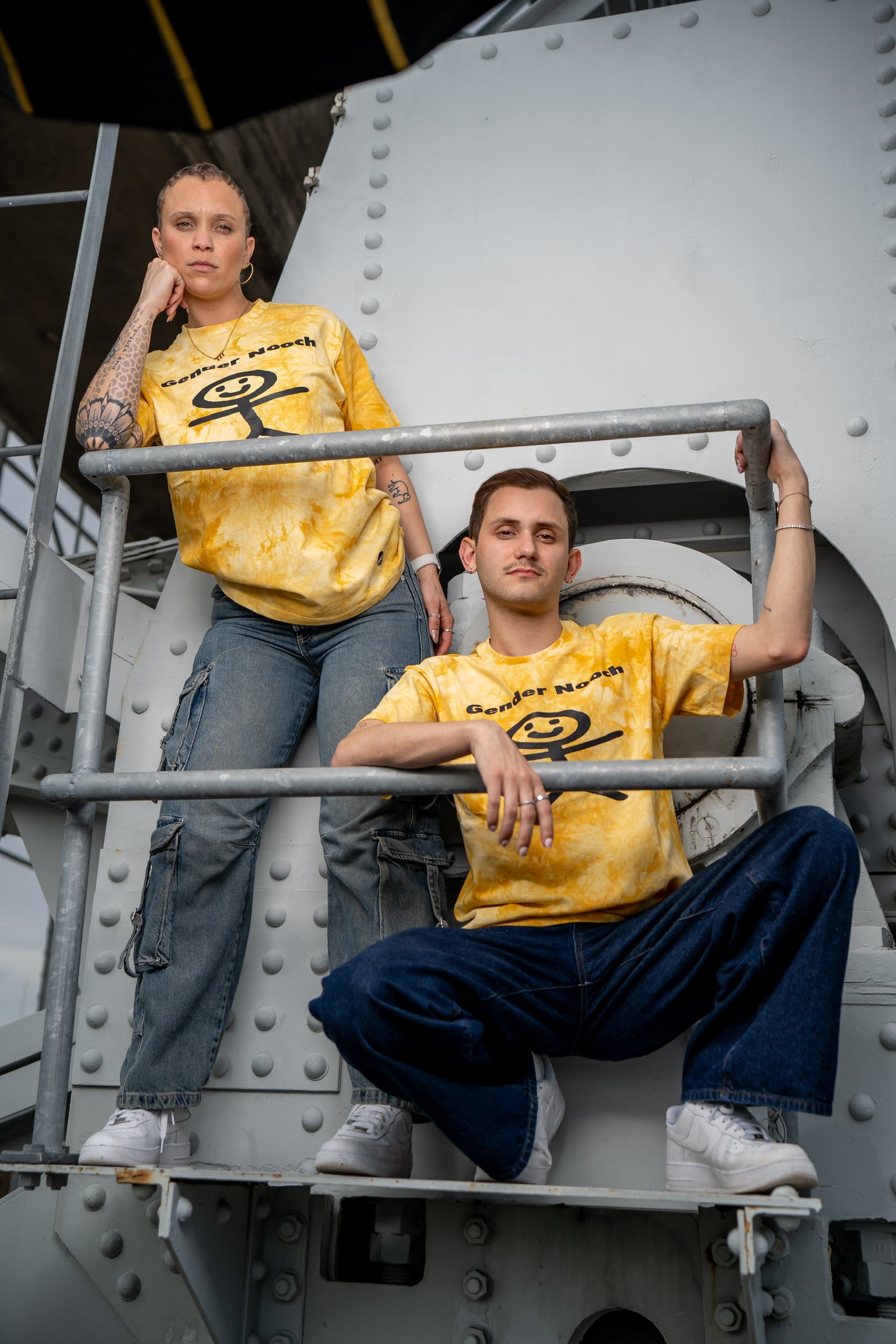 Two people, a man and a woman wearing the gender nooch tees by merakitay. Acid wash yellow gender-neutral tee shirt. Embroidered logo and screen printed. crew neck with tighter neckline, and short sleeves. Unisex, nonbinary clothing. 