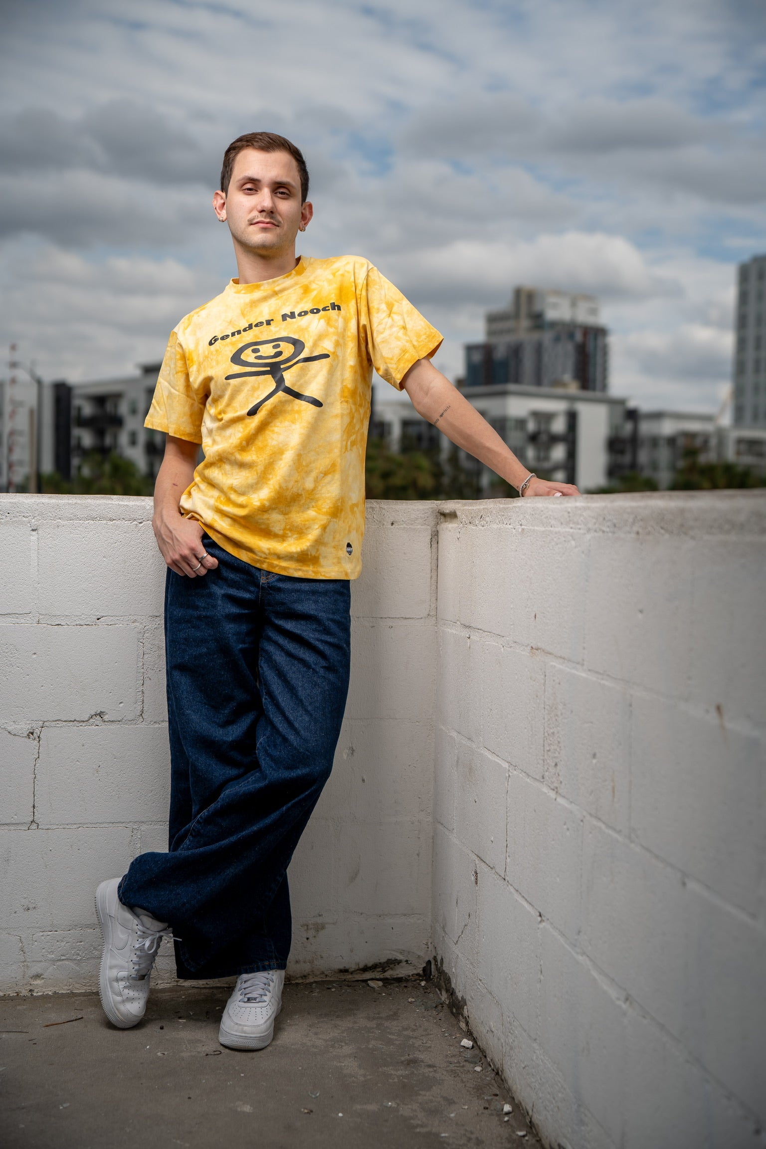 A man wearing the gender nooch merakitay tee shirt. Acid wash yellow gender-neutral tee shirt. Embroidered logo and screen printed. crew neck with tighter neckline, and short sleeves. Unisex, nonbinary clothing. 