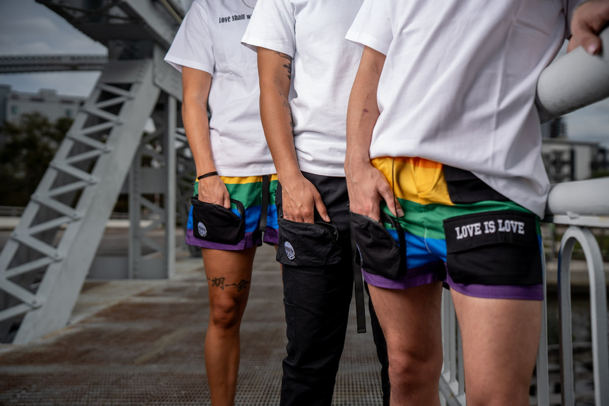 Emphasis on merakitay rainbow pride cargo shorts and merakitay black cargo pants. Hands in one of the six pockets. Gender neutral cargo shorts. Rainbow in color, embroidered logo and words, adjustable waistband, includes belt, and has six (6) pockets. Mid-thigh length, 3-5 inch inseam. Unisex shorts. LGBTQ+ pride cargo shorts perfect for parades. 