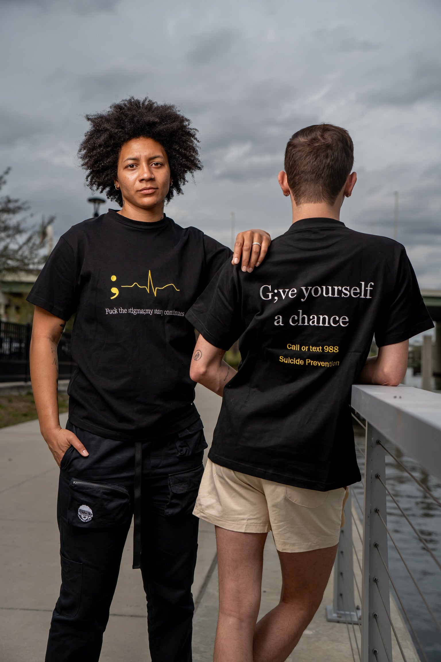 Woman and man wearing merakitay mental health tee. All embroidered mental health merakitay tee shirt. Stigma tee. Semi colon tee. Fuck the stigma, my story continues tee. Suicide prevention hotline tee. Give yourself a chance.  Embroidered logo. Black with yellow and white embroidery.